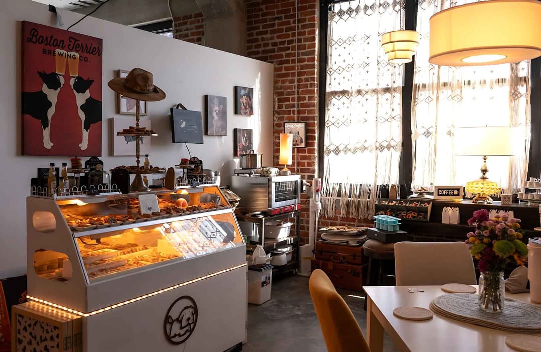 A cozy bakery and retail space with a glass display case full of baked goods, decorative items, and dog-themed art on the walls. A warm, inviting atmosphere with brick walls, soft lighting, and rustic decor.