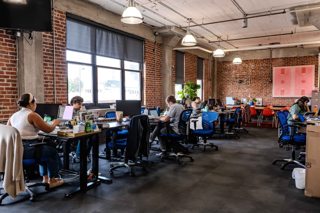 A vibrant coworking office space in Kansas City, featuring exposed brick walls, rows of desks with professionals working on laptops, and bright natural light from large windows. The industrial aesthetic includes plants, artwork, and modern furnishings.