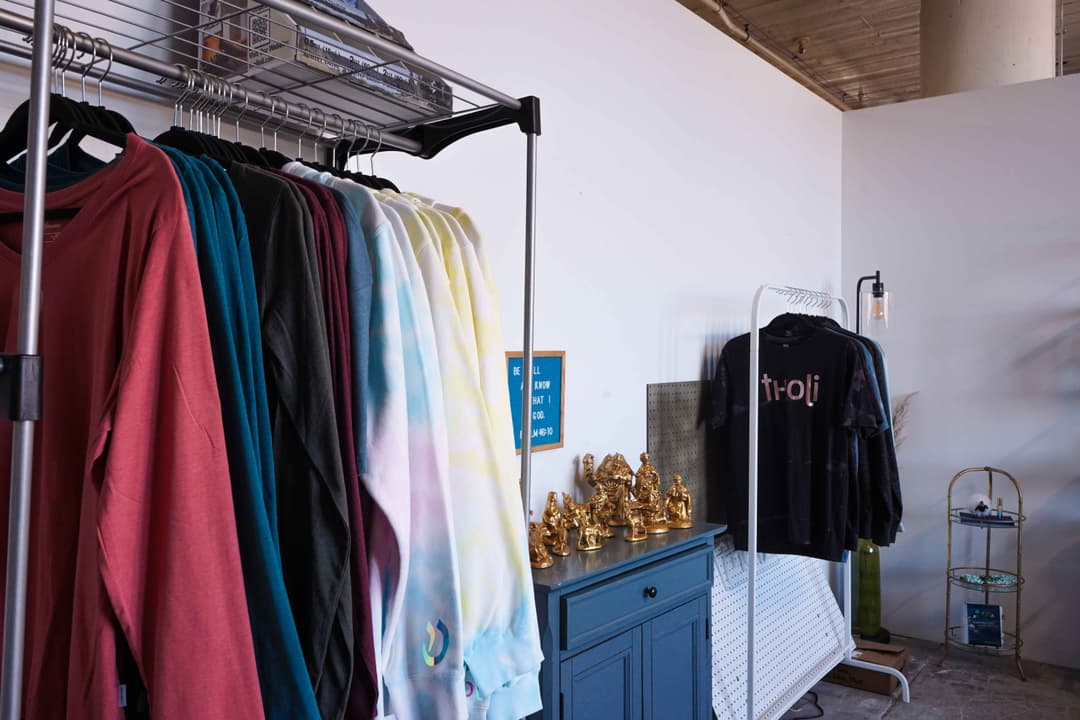 A retail and workspace featuring colorful clothing racks, a blue cabinet displaying golden figurines, and a modern, minimalist setup with a white pegboard and industrial lighting. The space has a clean, organized design with a creative flair.