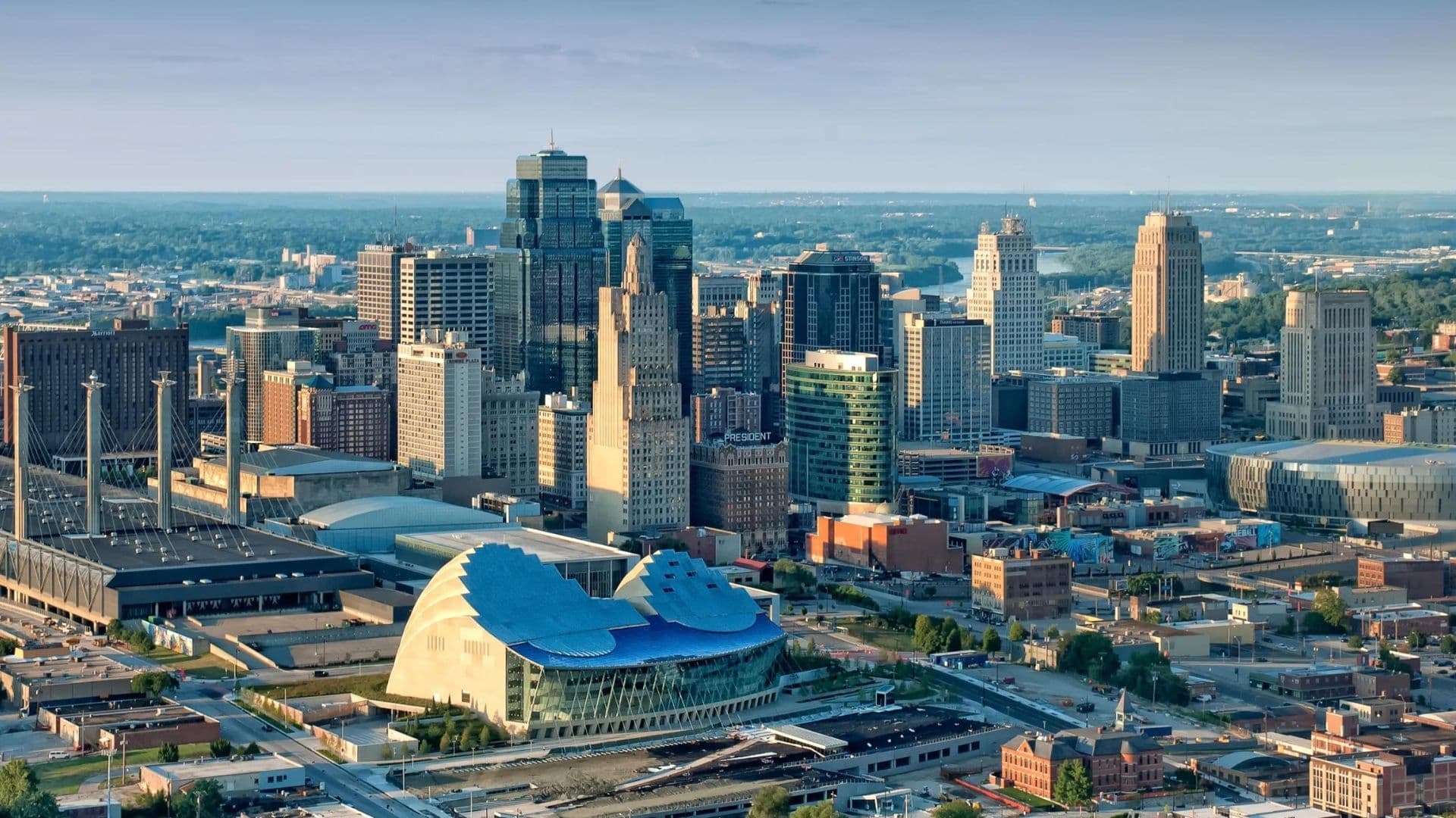 A stunning aerial view of the Kansas City, Missouri skyline featuring prominent skyscrapers, historic buildings, and the Kauffman Center for the Performing Arts with its iconic blue roof in the foreground. This image represents the vibrant city where Elevator offers co-warehousing, office space, and coworking solutions.